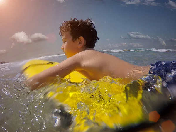 Tini fiú szörfözik a trópusi tengerparton Ázsiában. Egy gyerek szörfdeszkán az óceán hullámain. Aktív vízi sportok gyerekeknek. A srác úszik a deszkával. Fiatal szörfös az egzotikus tengeren. Úszás és szemviselet gyerekeknek. — Stock Fotó