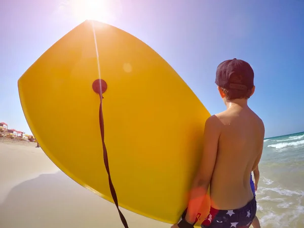 Menino feliz em boné em calções de banho fica na praia na areia dourada e detém uma prancha de natação, prancha de boogie de cor amarela contra o céu azul e o mar com pequenas ondas ao sol ensolarado . — Fotografia de Stock