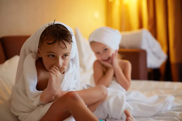 Happy laughing kids, boy and girl in soft bathrobe after bath play on white bed with white pillows in sunny bedroom. Child in clean and dry towel. Wash, infant hygiene, health and skin care. — Stock Photo, Image