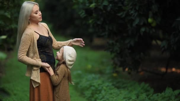 Zijaanzicht van liefdevolle moeder met meisje op groene weide in groene zonnige boomgaard — Stockvideo