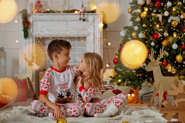 Happy little kids in matching red and green striped pajamas decorate Christmas tree in beautiful living room with traditional fire place. Children opening presents on Xmas eve.