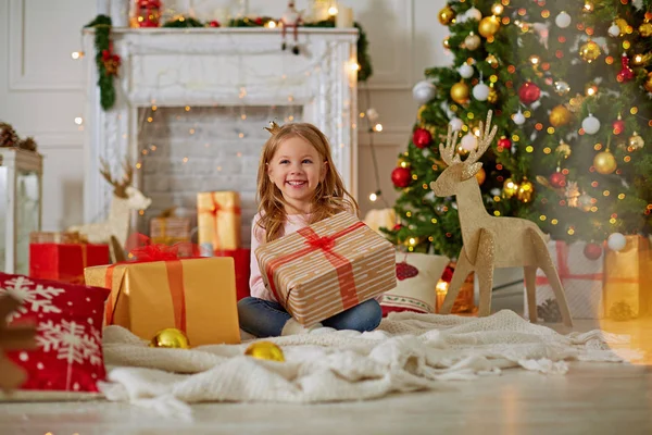 Entusiasmada menina sorrindo, abrindo presentes de Natal. Belamente decorado árvore de Natal e casa com luzes e lanterna . — Fotografia de Stock