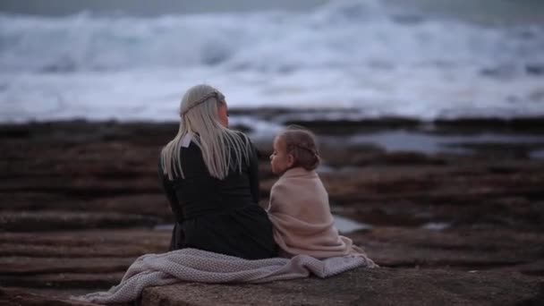 Vista trasera de una mujer rubia con una niña acurrucada a cuadros en la costa rocosa en el crepúsculo — Vídeo de stock