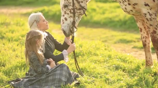 緑の牧草地に斑点を付けられた馬をなでる、ドレスの少女と若い母親の正面図 — ストック動画