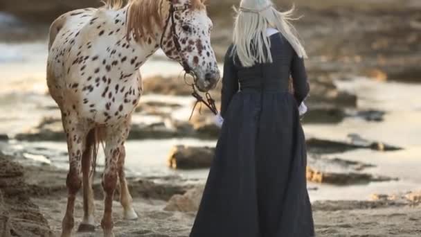 Therapie met paarden. Moeder en dochter in elegante jurk lopen op rotsachtige kust met mooie vlekkerige paard — Stockvideo