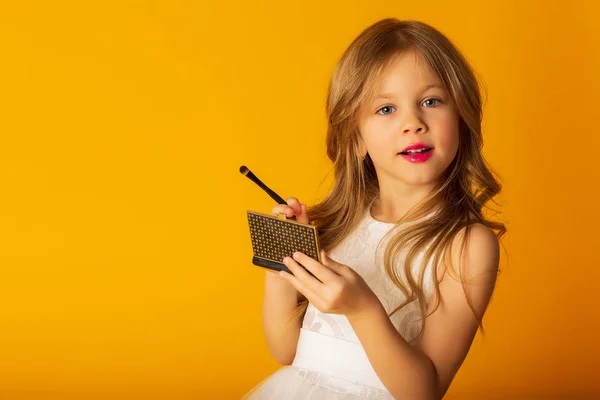 Adorable niña sosteniendo el espejo de bolsillo y aplicando maquillaje mientras está de pie sobre fondo amarillo — Foto de Stock