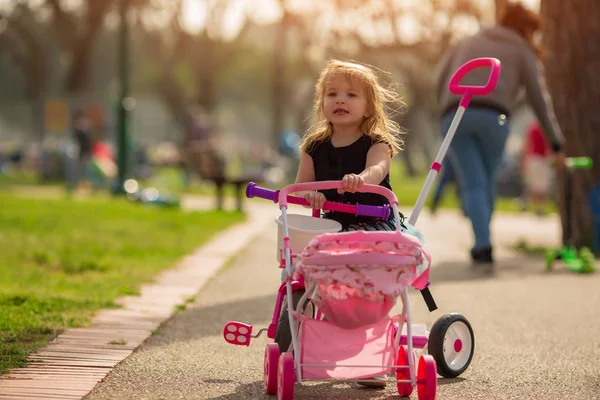 Hermosa niña está jugando con una muñeca en un cochecito al aire libre al atardecer del día — Foto de Stock