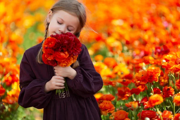 Bedårande liten flicka med blont hår i en enkel klänning som håller bukett med Butter cups och lukta dem med ögonen stängda i en äng med blommor på landsbygden — Stockfoto