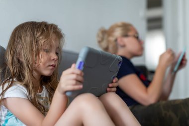 Kızlar kanepede oturup kitap okuyorlar. Kız elinde telefon varken kadın tablet tutar