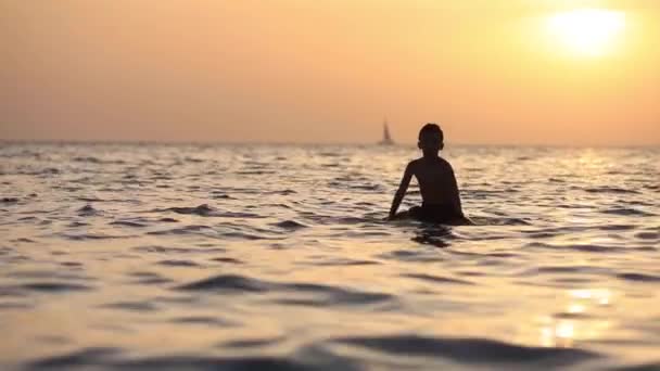 Silueta de un niño flotando en el mar con tabla de surf sobre el excelente fondo de la puesta de sol naranja — Vídeos de Stock