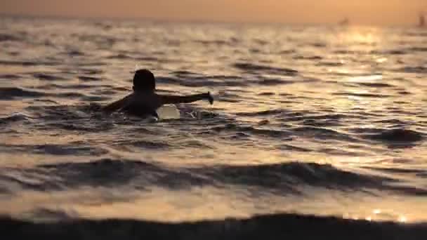 Niño flotando en el mar con una tabla de surf en el excelente fondo de naranja puesta de sol — Vídeos de Stock