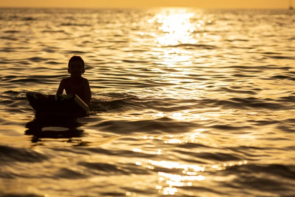 Silhueta menino sozinho em uma placa no mar ao pôr do sol — Fotografia de Stock