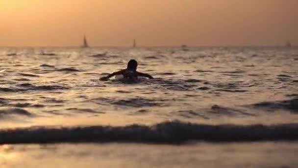 Silhueta de um menino flutuando no mar com prancha de surf no excelente fundo do pôr-do-sol laranja — Vídeo de Stock