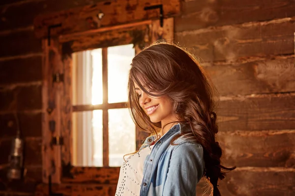 Młoda kobieta z długimi włosami uśmiechnięta w pobliżu okna w wiejskim domu. — Zdjęcie stockowe