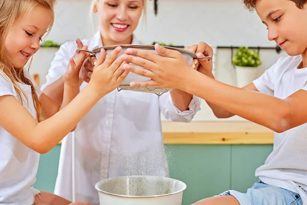 Madre feliz y niños pequeños con tamizar la harina y la masa para hornear en la cocina casera . — Foto de Stock
