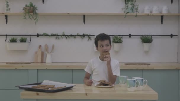 Garoto tomar café da manhã na cozinha moderna. O rapaz come bolachas com leite. Mesa branca na cozinha — Vídeo de Stock