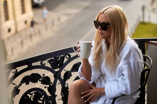 Schöne sexy Blondine im weißen Bademantel und Sonnenbrille sitzt mit einer Tasse Kaffee auf dem Balkon — Stockfoto