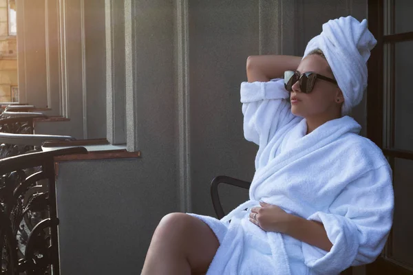 Giovane donna in accappatoio bianco, asciugamano per capelli e occhiali da sole seduta su una sedia sulla terrazza balcone — Foto Stock