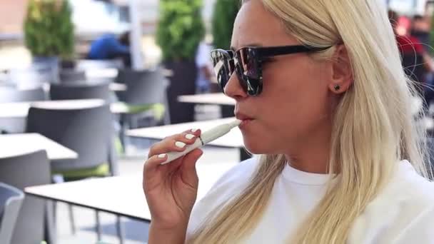 Schöne Blondine raucht elektronische Zigarette auf der Sommerterrasse des Restaurants — Stockvideo