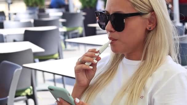 Primer plano de una rubia en gafas de sol con un teléfono y un cigarrillo electrónico en un café al aire libre — Vídeo de stock