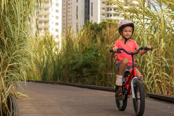 Маленька дівчинка їде на велосипедах вздовж дерев'яної доріжки за межами міста в шоломах як захисне обладнання — стокове фото