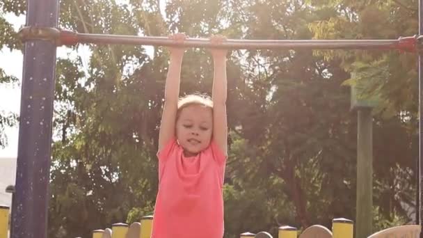 Милая маленькая девочка играет на детской площадке и висит на горизонтальной панели . — стоковое видео