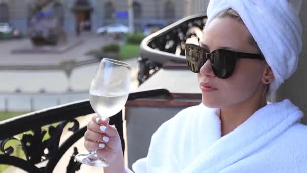Menina elegante em um roupão de banho branco e uma toalha branca na cabeça, desfrutando de um copo de vinho branco — Vídeo de Stock