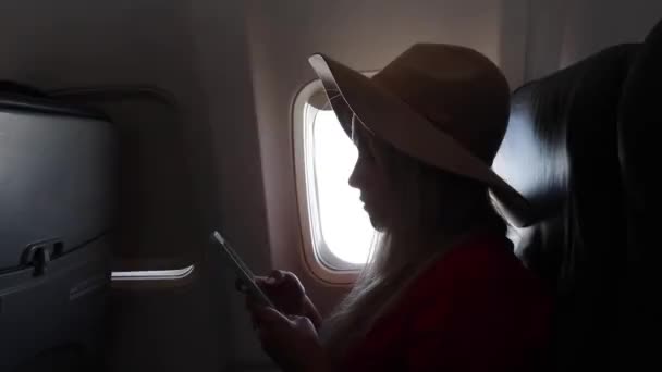 Mujer turista en un sombrero se sienta cerca de la ventana de un avión al atardecer y usando un teléfono móvil mientras vuela — Vídeo de stock