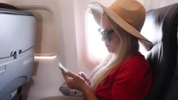 Turist i en hatt sitter nära fönstret på ett flygplan vid solnedgången och använder en mobiltelefon medan du flyger — Stockvideo