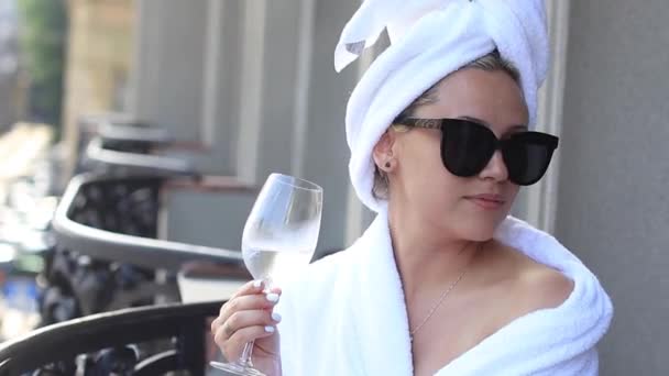 Linda loira em um casaco branco desfrutando de um copo de vinho branco sentado em um terraço do hotel contra — Vídeo de Stock