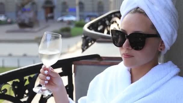 Menina em um roupão de banho branco e uma toalha na cabeça, desfrutando de um copo de vinho branco — Vídeo de Stock