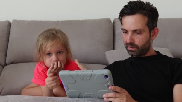 Padre e hija sentados en un sofá gris y usando una tableta digital — Vídeo de stock