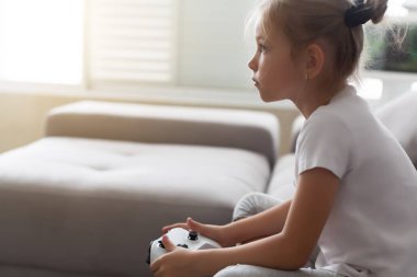 Zevkli bir çocuk, günlük kıyafetleriyle oturma odasındaki kanepede otururken joystickle oynuyor.