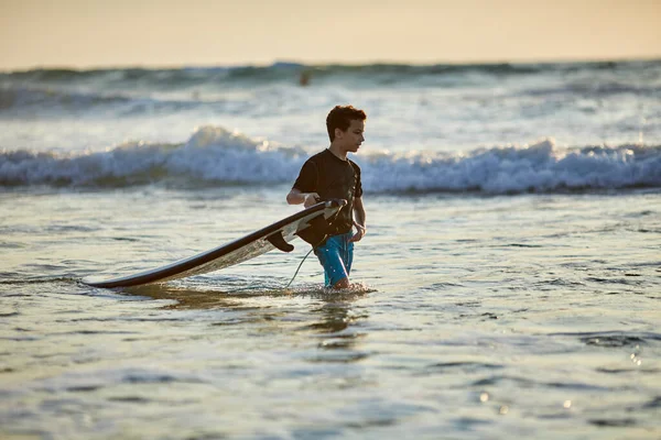 Vista lateral de criança destemida flutuando prancha de surf no oceano com ondas na noite ensolarada — Fotografia de Stock