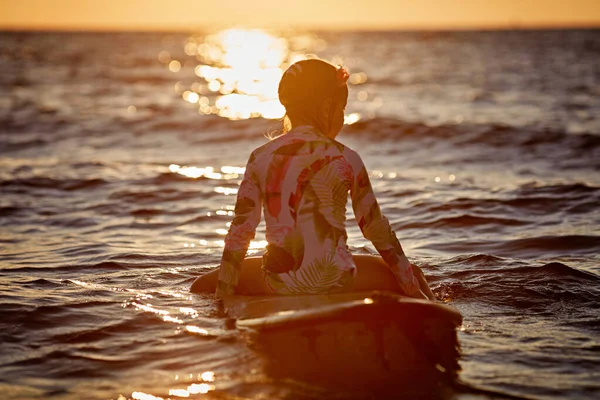 Urlaub am Strand. Entspanntes kleines Mädchen sitzt mit Surfbrett auf dem Wasser und genießt den Sonnenuntergang — Stockfoto