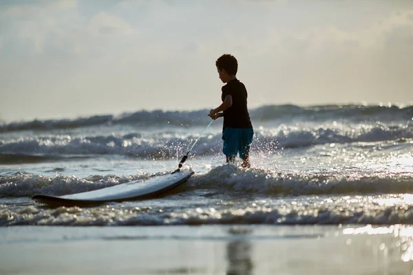 Кавказский мальчик прогуливается по пляжу с белой доской для серфинга, голубой океан на заднем плане — стоковое фото