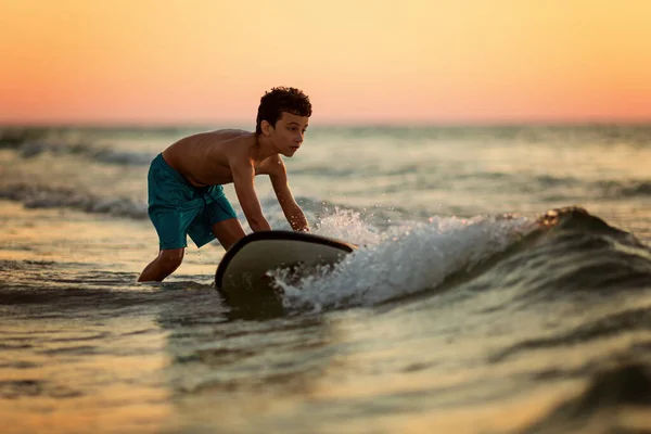 Vista lateral de niño intrépido tabla de surf flotante en el océano con olas en la tarde soleada —  Fotos de Stock