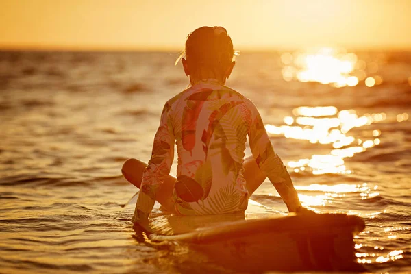 Chica vista trasera sentada en una tabla de surf durante el atardecer, mar tranquilo sin olas de luz naranja —  Fotos de Stock