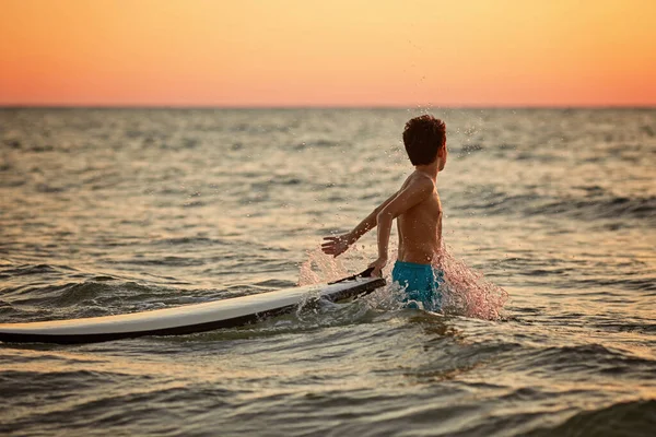 Menino surfista feliz faz salpicos com as mãos no pôr-do-sol dourado — Fotografia de Stock