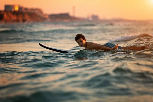 Vista lateral de niño intrépido tabla de surf flotante en el océano con olas en la tarde soleada —  Fotos de Stock