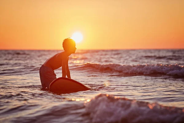 Vista lateral de criança destemida flutuando prancha de surf no oceano com ondas na noite ensolarada — Fotografia de Stock