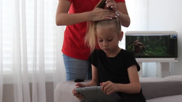 Mother combs cute daughter hair, which looks at tablet. Mother spends time with daughter — Stock Video