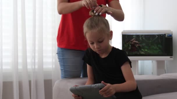 Η μητέρα χτενίζει τα χαριτωμένα μαλλιά της κόρης της, η οποία κοιτάζει ταμπλέτα. Η μητέρα περνάει χρόνο με την κόρη. — Αρχείο Βίντεο
