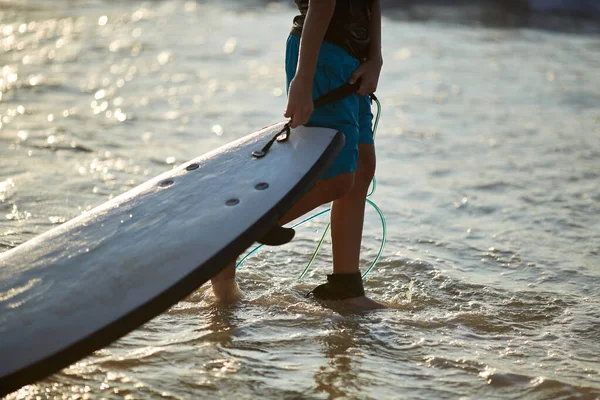Um rapaz surfista corre no oceano com prancha de surf. Imagem de close-up salpicos de água e pernas, luz do pôr do sol — Fotografia de Stock