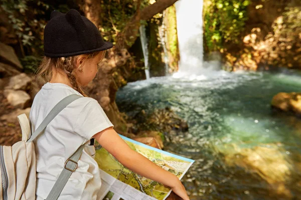 이 어린 소녀는 폭포 근처의 화창 한 날 숲 속에서 독서 지도를 보고 있다 — 스톡 사진