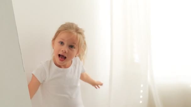 Schattig klein meisje verstopt achter een gordijn, kijken uit en plezier hebben. Gelukkig kind spelen verbergen en zoeken. — Stockvideo