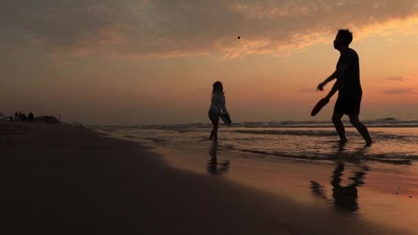 Aktív fiatal apa és lánya játszik strandtenisz játék naplemente fény. Nyaralás táj kilátás nap, tengeri hullámok és hegyek a horizonton. Boldog férfi és lány sziluettek a szerelem sport nyár — Stock videók