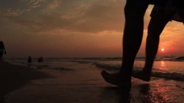 Yaşlı çift gün batımında el ele tutuşup sahilde yürür ve ayaklarını ıslatırlar.