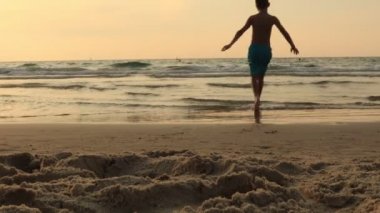 Erkek çocuk gün batımında bir deniz sahilinde zıplıyor. Hiperaktif çocuk koş ve kumsalda gülümse..