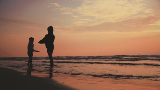 アクティブな子供たちは日没の光の中でビーチテニスゲームをプレイ。水平線上の太陽、海の波と山と休暇の風景ビュー。ハッピー男と女の子シルエットで愛スポーツ夏 — ストック動画